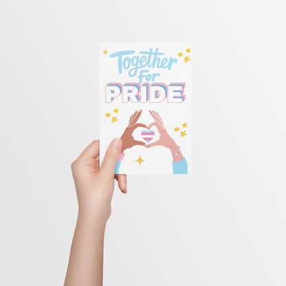 A hand holding a Trans Pride celebration card with bold text in trans flag colors reading “Together for Pride.” Features a heart in trans flag colors formed by two hands in a heart shape. Designed to celebrate Transgender Pride with allies, family and friends. 