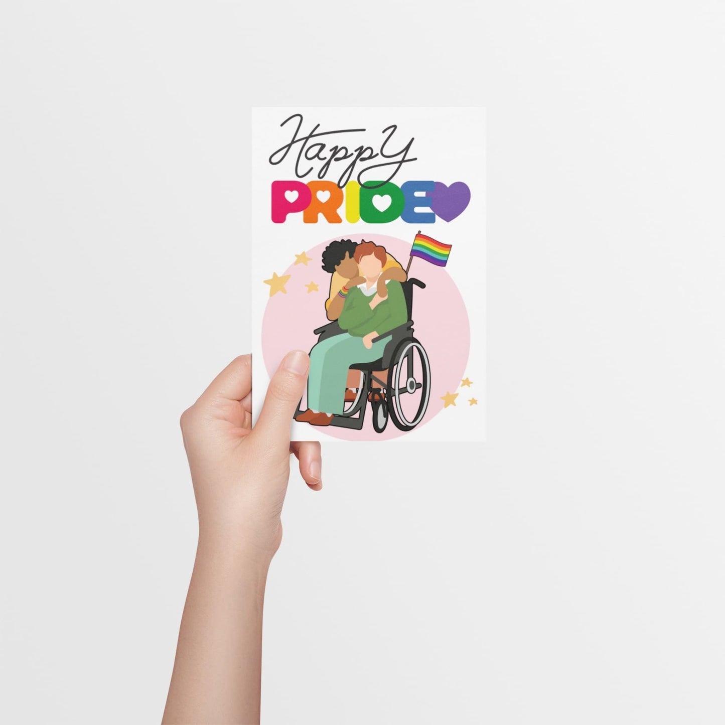 A hand holding a card with rainbow letters spelling the word 'Pride, with queer couple on the front using a wheelchair and a LGBTQ+ rainbow flag. This card is designed to celebrate and represent disabled and queer people on Pride. 